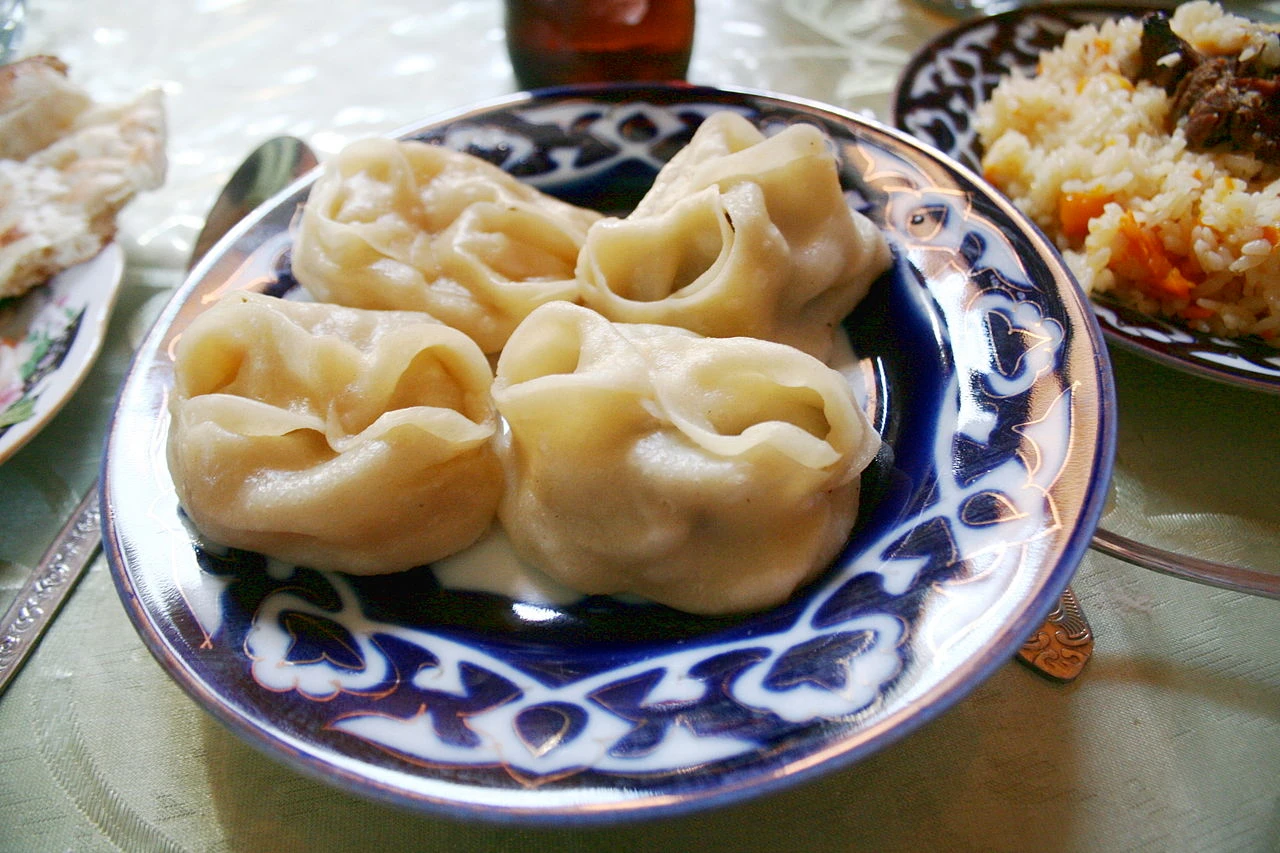 Manti uzbeki appena cotti al vapore, su un piatto decorato, con una ciotola di qatiq a lato